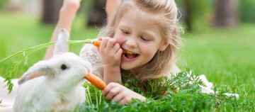 Glückliches blondes Mädchen teilt eine Karotte mit einem weißen Kaninchen im grünen Gras im Freien