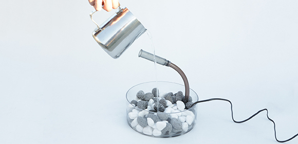 Schritt 5 Moderner Luftbefeuchter mit schwarzen und weißen Steinen in einem transparenten Behälter, Wasser wird aus einem Metallkrug gegossen, minimalistisches Design.