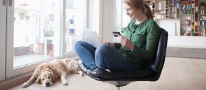 Jeune femme assise sur une chaise moderne effectuant des achats en ligne avec une carte de crédit et un ordinateur portable, un golden retriever détendu à ses côtés.