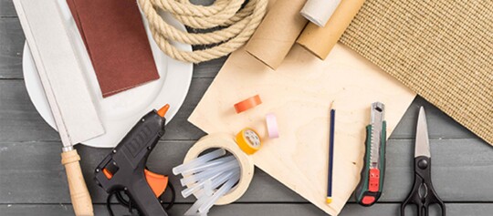 Verschiedene Bastel- und Heimwerkerwerkzeuge wie Handsäge, Heißklebepistole, Klebebänder, Seil, Cutter, Schere und Bleistift auf einem Holztisch