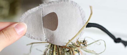 Nahaufnahme einer Hand, die ein weißes, ovales Stoffobjekt mit braunem Fleck hält, im Hintergrund ein Bündel trockenen Grases, das mit einer schwarzen Schnur gebunden ist.