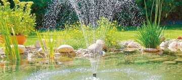 Gartenteich mit sprudelndem Wasserbrunnen, umgeben von grünen Pflanzen, Steinen und blühendem Gras