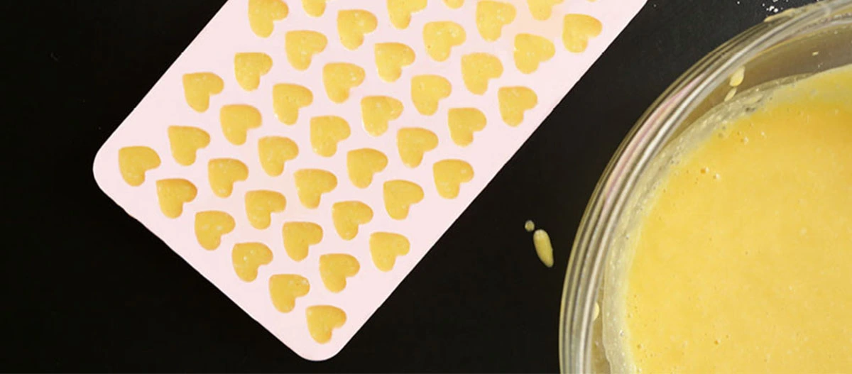 Silikonform mit kleinen herzförmigen Teigstücken und Schüssel mit gelber Mischung auf schwarzem Hintergrund