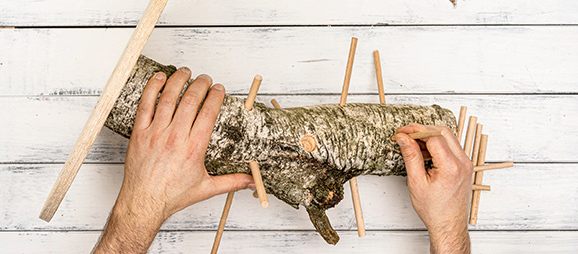 DIY_Futterbaum_Beitrag_05_1200x527 Hände stecken Holzstäbe in einen Baumstamm aus Birkenholz auf einem weißen Holztisch – Holzhandwerk DIY-Projekt