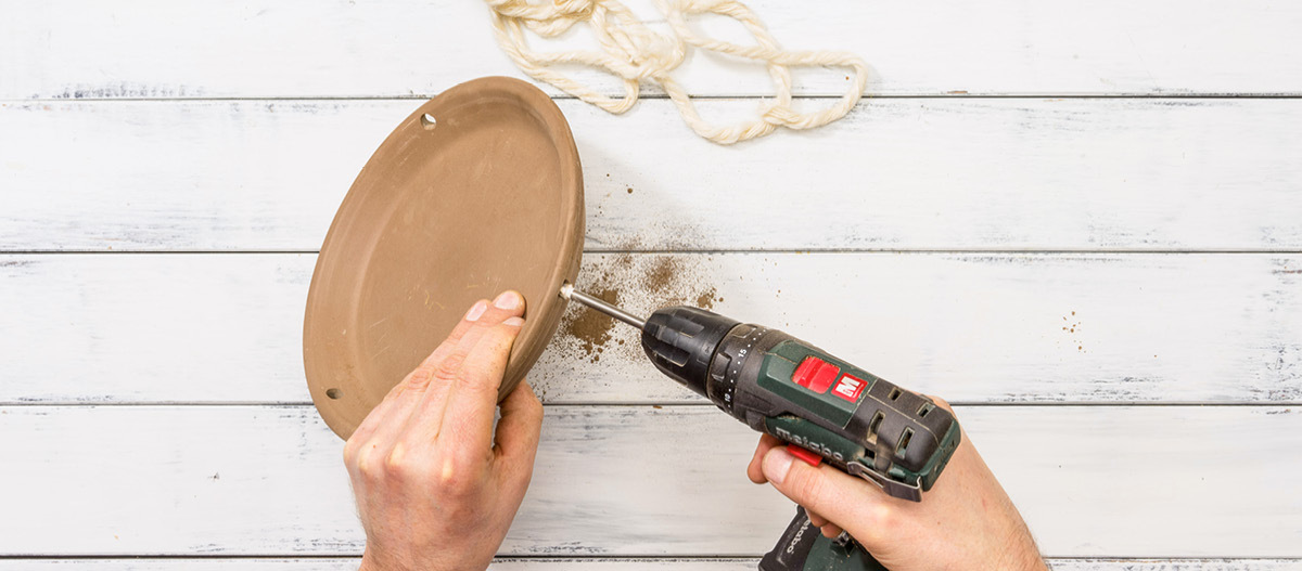 Schritt 1 Person bohrt Loch in braunen Terrakotta-Untersetzer mit Akku-Bohrmaschine auf weißem Holzhintergrund