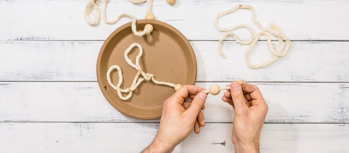 Schritt 2 Hände fädeln große Holzperlen auf eine dicke cremefarbene Schnur, die an einem braunen Keramikteller befestigt ist, auf weißem Holzhintergrund.