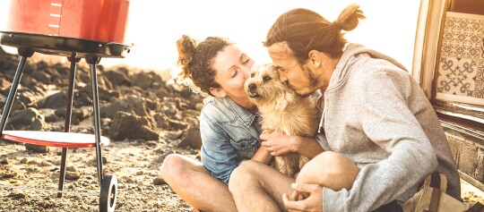 Glückliches Paar küsst kleinen Hund beim Camping neben rotem tragbarem Grill im Sonnenlicht