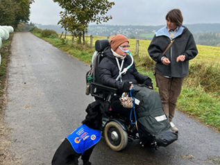 Person im Elektrorollstuhl mit Assistenzhund auf Landstraße, begleitet von einer weiteren Person, ländliche Umgebung mit grünen Feldern und Bäumen