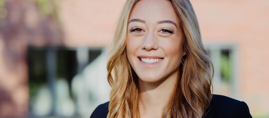 Porträt einer jungen, lächelnden Frau mit langen blonden Haaren und dunklem Blazer im Freien vor unscharfem Hintergrund.