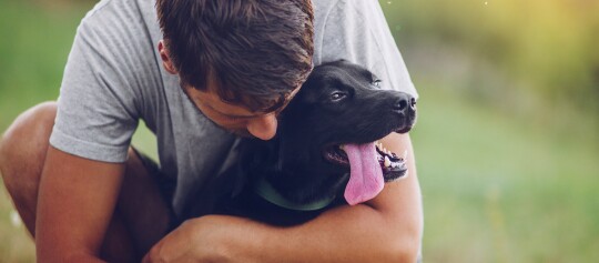 Mann umarmt glücklichen schwarzen Hund mit herausgestreckter Zunge im Freien