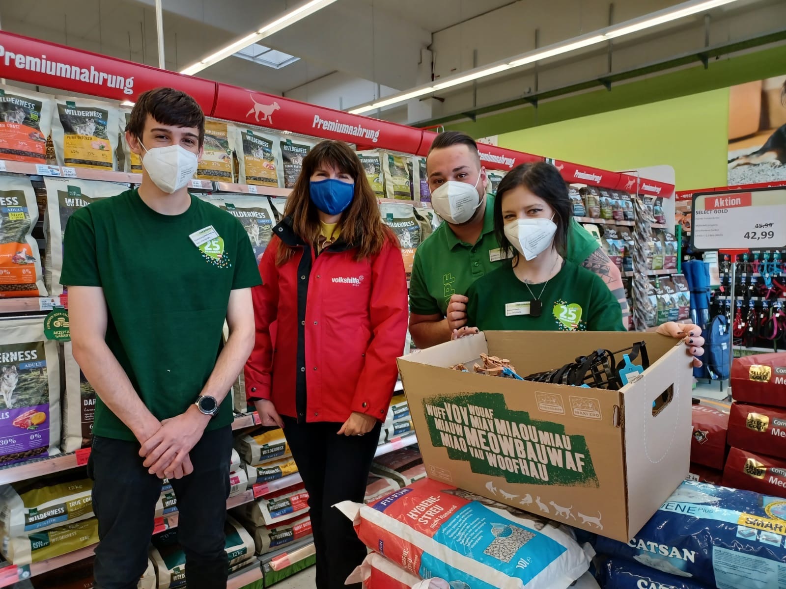 Vier Mitarbeiter in einem Tierfachgeschäft mit Premium-Tiernahrung und einer Spendenbox voller Haustierbedarf, alle mit Schutzmasken.