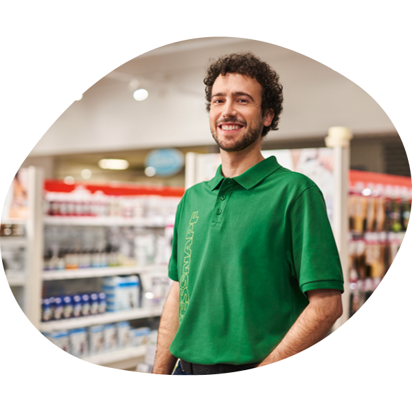 NE_KW8_ExpertCampaign_LB_NL3 Junger Mann mit grünem Poloshirt lächelt freundlich in einem Supermarkt mit vollen Regalen im Hintergrund.