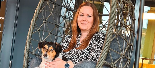 Frau mit langem, glattem, hellbraunem Haar sitzt in einem modernen, geflochtenen Hängesessel und hält einen kleinen Hund mit schwarzem, weißem und braunem Fell auf dem Schoß in einem gemütlichen, zeitgemäßen Wohnzimmer.