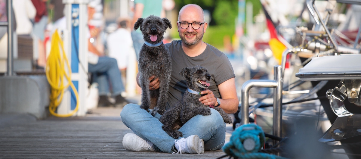 Glücklicher Mann sitzt am Hafensteg mit zwei kleinen schwarzen Hunden und lächelt in die Kamera
