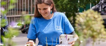 Junge Frau in blauem NABU-T-Shirt lächelt und hält ein Pflanzenbuch im Garten, umgeben von Blumen und Grün.