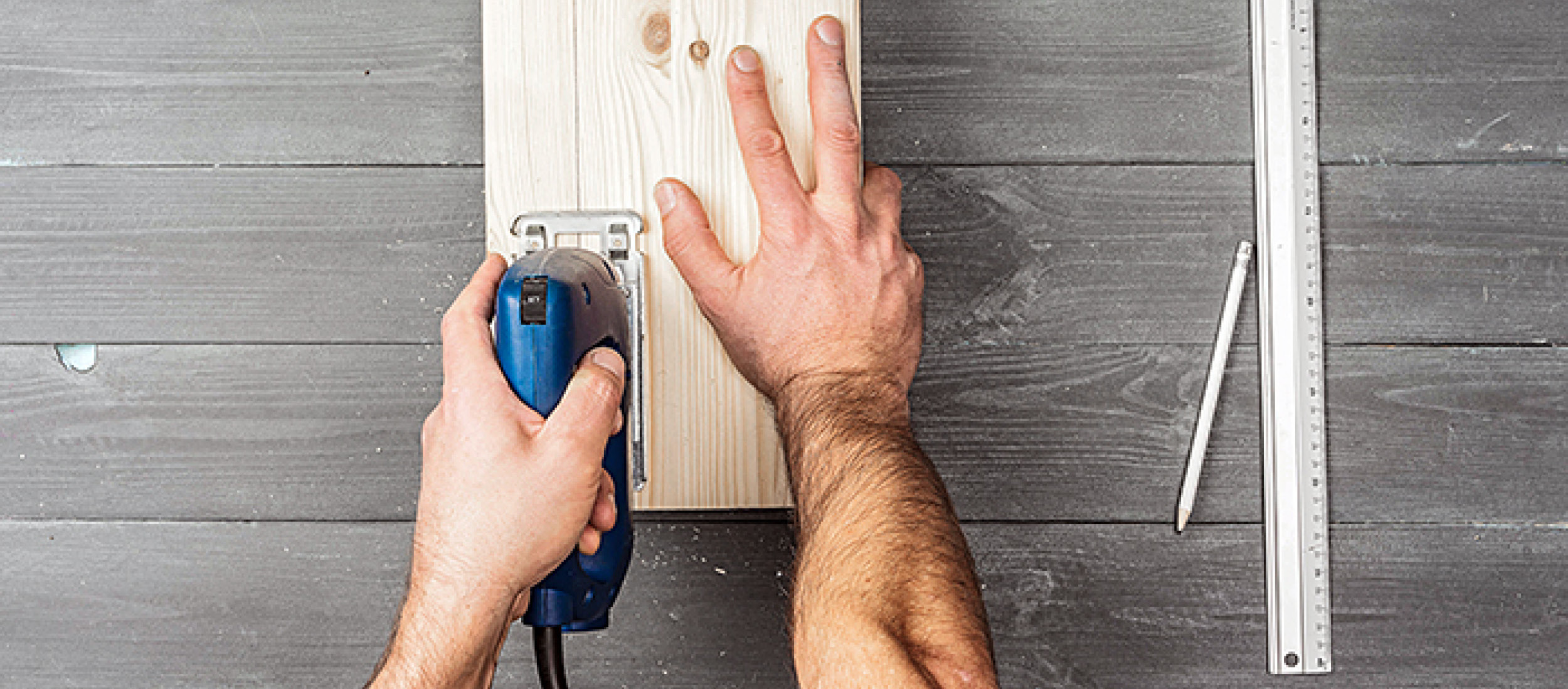 Person schneidet Holz mit elektrischer Stichsäge auf dunklem Holztisch mit Lineal und Bleistift