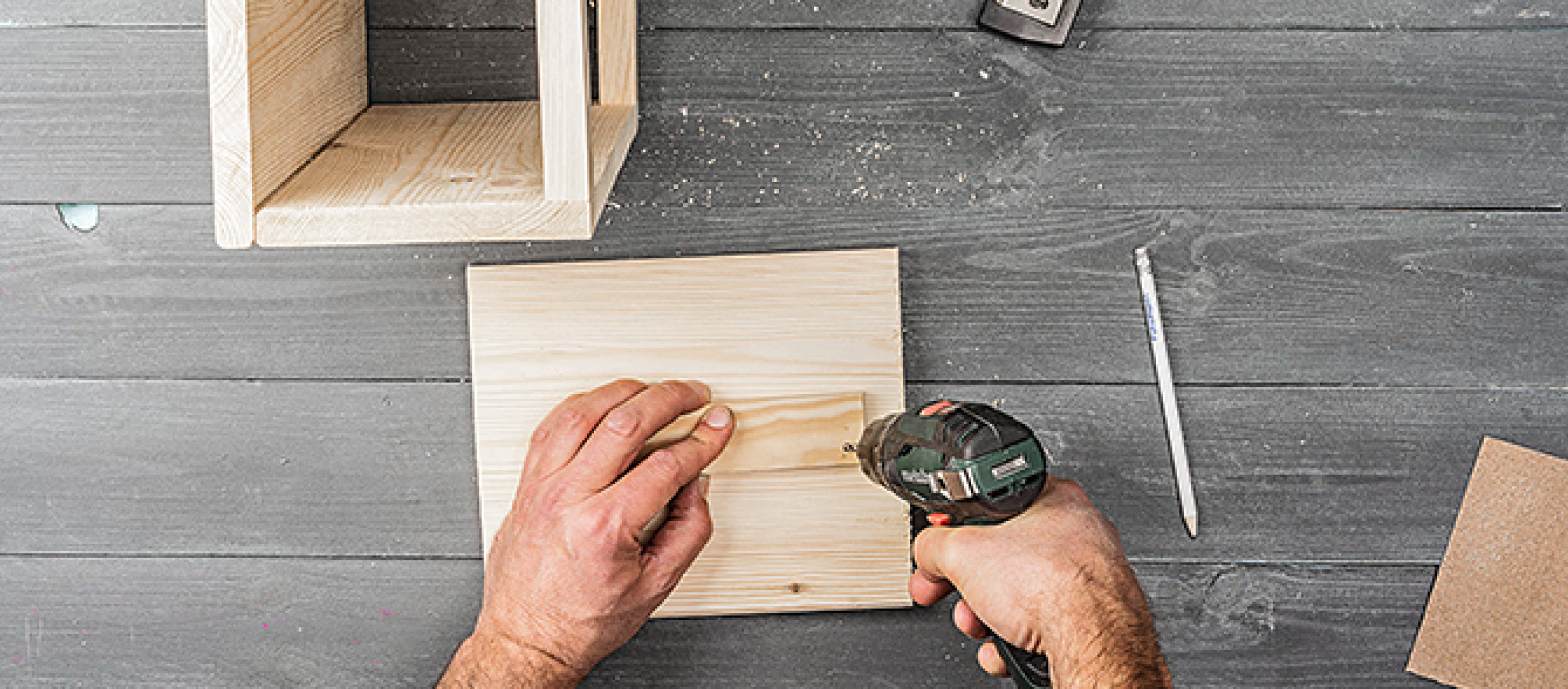 Mann benutzt Akkuschrauber, um ein kleines Holzstück auf einem Holzbrett bei einem DIY-Holzprojekt auf einem grauen Holztisch zu befestigen