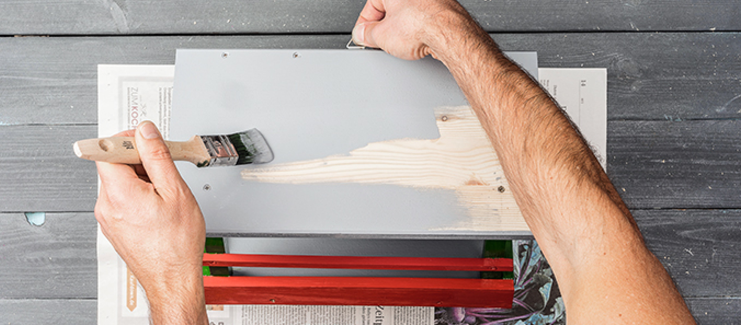 Person streicht Holzbrett mit grauer Farbe auf einem Tisch mit Pinsel und rotem Holzkasten