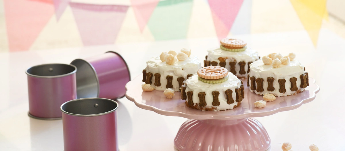 Petits gâteaux ronds pour chiens avec glaçage blanc, décorés de biscuits en forme d'os et de friandises pour chiens sur un présentoir à gâteau rose, à côté de moules à pâtisserie violets.