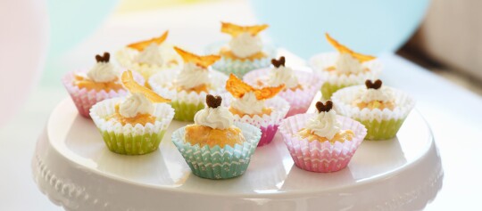 Kleine bunte Cupcakes mit weißer Creme und dekorativen Toppings auf einem weißen Kuchenständer, perfekt für Partys und Feiern.