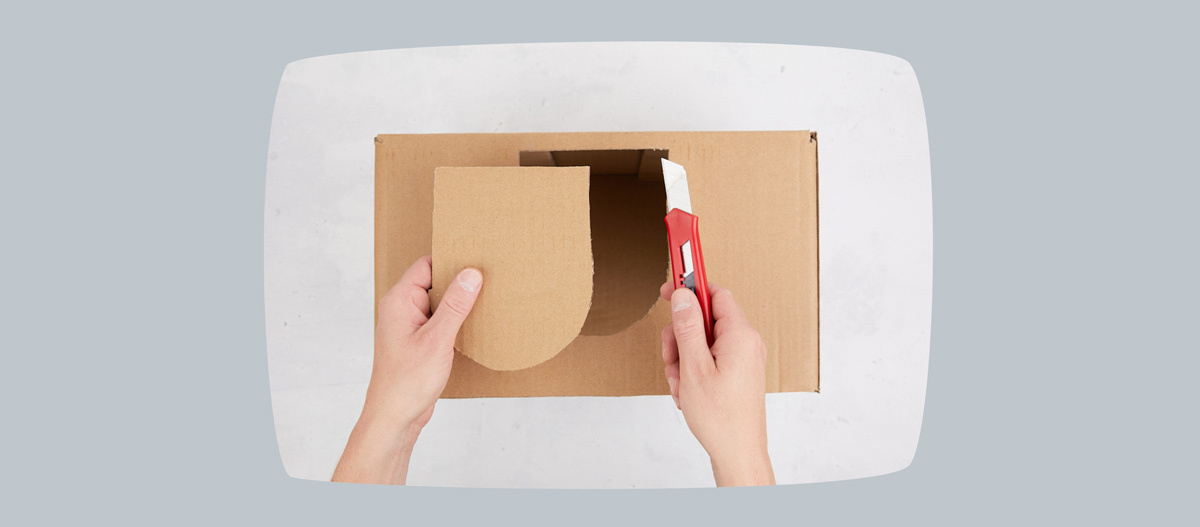 0622_Online_RATGEBER_1200x527px_DIY_Meerschweinchen_03 Hände schneiden Karton mit rotem Cuttermesser, Kartonstück wird ausgeschnitten