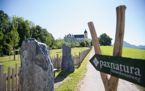 Weg mit großen Steinen und Schild 'paxnatura Naturbestattung' zu einem historischen Gebäude in grüner Landschaft
