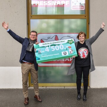 Deux personnes devant un magasin Fressnapf tiennent un grand chèque de don de 4 500 CHF pour Handicap Katzen Schweiz, les deux levant les bras en signe de célébration.