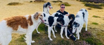 Mann sitzt im Freien mit fünf Borzoi-Hunden auf trockenem Gras und grüner Vegetation