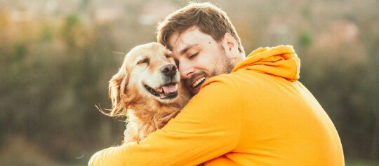 Glücklicher Mann in orangefarbenem Hoodie umarmt liebevoll seinen zufriedenen Golden Retriever im Freien