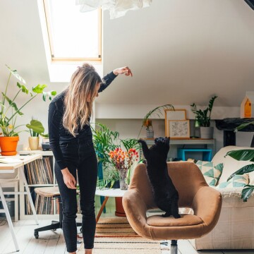 Femme jouant avec un chat noir sur un fauteuil marron dans un salon confortable avec de nombreuses plantes et lumière naturelle
