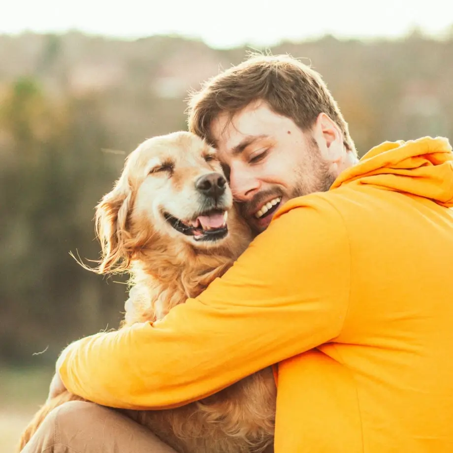 Glücklicher Mann in orangefarbenem Hoodie umarmt liebevoll seinen zufriedenen Golden Retriever im Freien