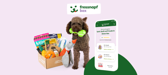 Brauner Pudel mit grünem Spielzeug neben geöffneter Fressnapf Box voller Haustierprodukte und Smartphone mit Fressnapf Box App