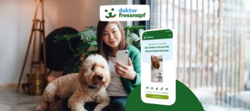 Junge Frau nutzt die Doktor Fressnapf App auf ihrem Smartphone, während ihr großer Hund entspannt neben ihr liegt – Online-Tierarztservice für Haustiere.