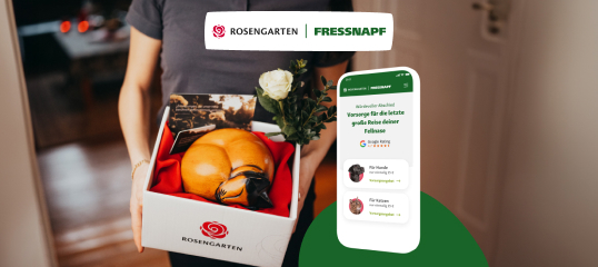 Person hält eine weiße Rosengarten-Box mit Kürbissen und einer weißen Rose, daneben ein Smartphone mit der Fressnapf-App zur Vorsorge für Haustiere.