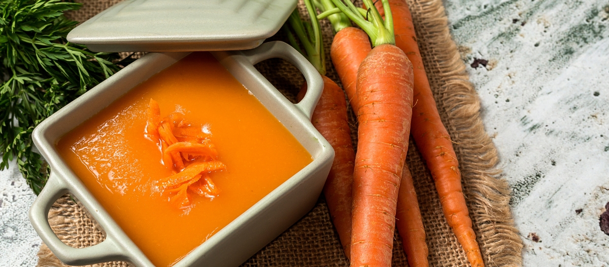 Frische Karottensuppe in einem quadratischen Keramikschälchen, garniert mit Karottenstreifen, neben frischen Karotten auf rustikalem Leinen.
