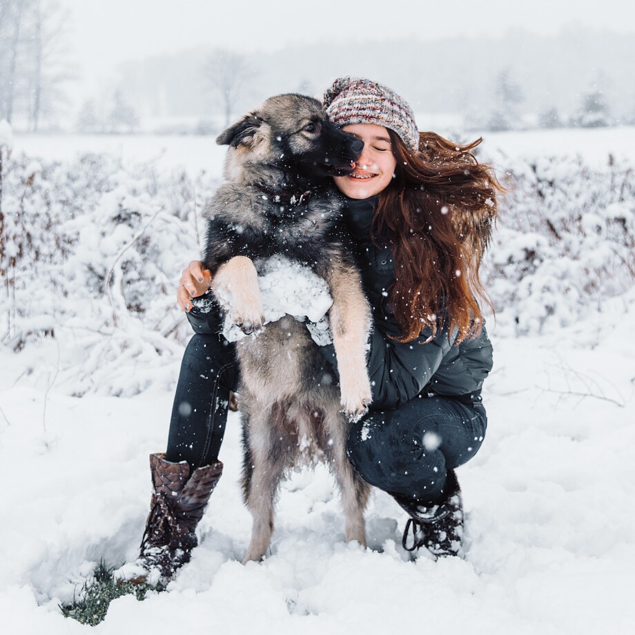 0124_APP_1500x1500px_09_Positiv_ins_neue_Jahr_Hund Junge Frau umarmt ihren großen Hund im verschneiten Winterwald, während Schneeflocken fallen.