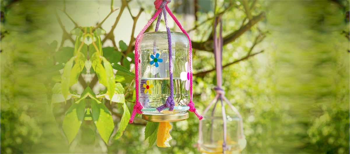 Selbstgemachter Vogelfutterspender aus einem umgedrehten Glas mit bunten Blumenaufklebern, hängend an einem Baumzweig im grünen Garten.