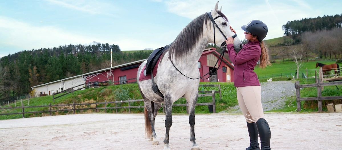 Frau in Reitkleidung mit Helm streichelt ein gesatteltes graues Pferd auf einem Reitplatz vor ländlicher Kulisse mit Scheune und grünen Hügeln.