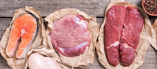 Verschiedene rohe Fleischsorten auf braunem Papier – Lachssteak, Hähnchenbrust, Schweinefleisch und Rindersteak mit Pfefferkörnern auf Holzuntergrund