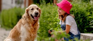 Glückliches Mädchen mit pinkem Hut spielt draußen mit einem freundlichen Golden Retriever im Garten.