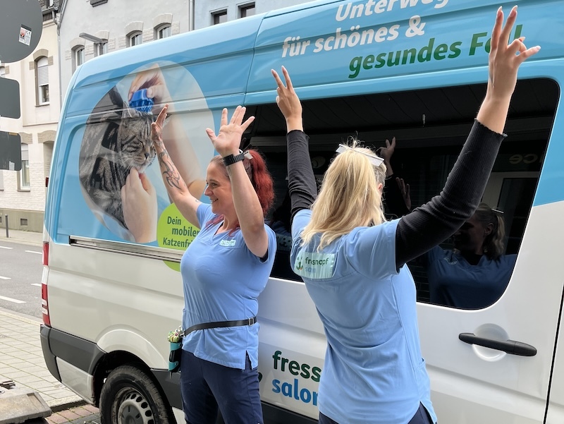 Zwei Frauen in blauen Shirts vor einem mobilen Katzenfriseur-Van mit erhobenen Armen, der für schönes und gesundes Fell wirbt.