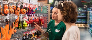 Deux femmes dans un magasin pour animaux examinent et discutent une sélection de jouets pour chiens accrochés au mur.