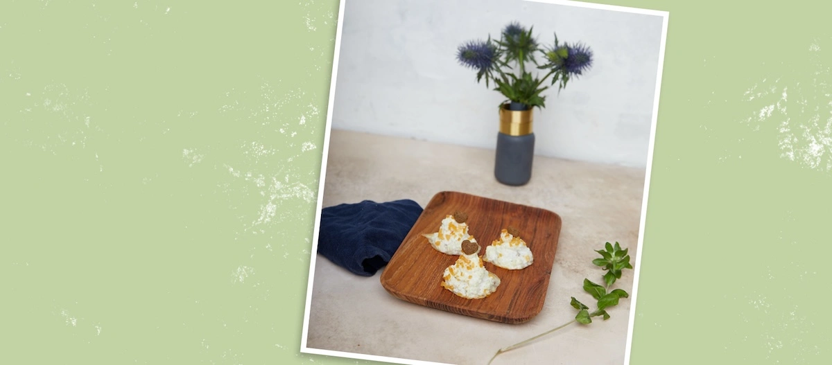 Drei gebackene Baiserhäufchen mit brauner Garnitur auf einem Holztablett, dekoriert mit lila Distelblumen in einer Vase und einem grünen Blatt