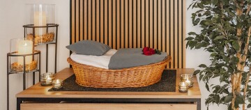 Gemütlicher Korb mit Kissen und Decke auf Holzkommode, umgeben von brennenden Kerzen und grüner Pflanze, dekoratives Schlafzimmer-Setup.