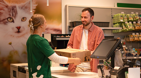 Servicewelt_CC Freundlicher Kunde übergibt Kassiererin im Zoofachgeschäft eine Kartonbox mit Katzenfutter
