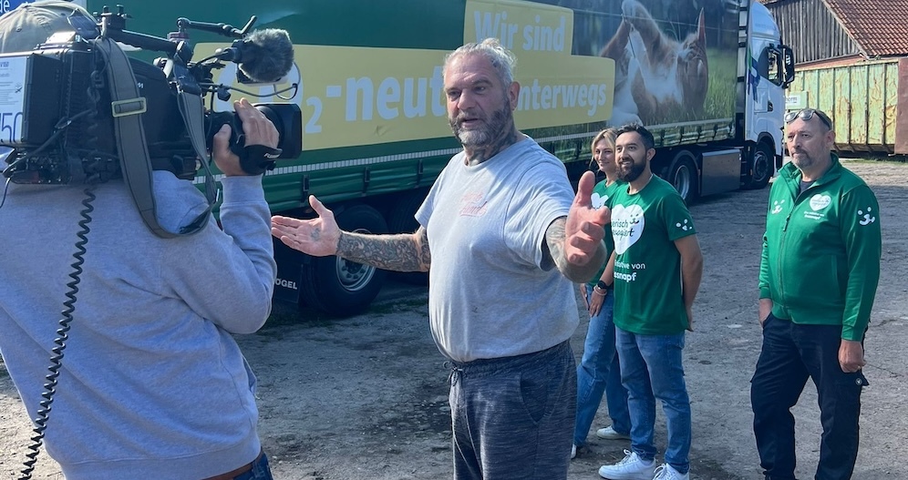 Mann mit Tattoos spricht vor der Kamera, während drei Personen in grünen Shirts zuschauen, vor einem grünen LKW mit der Aufschrift „Wir sind H2-neutral unterwegs“.