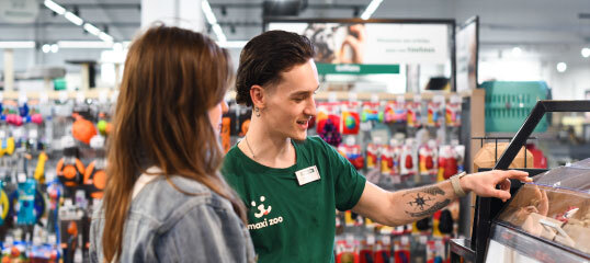 Employé en t-shirt vert Maxi Zoo conseille une cliente devant une vitrine en verre avec des produits pour animaux.