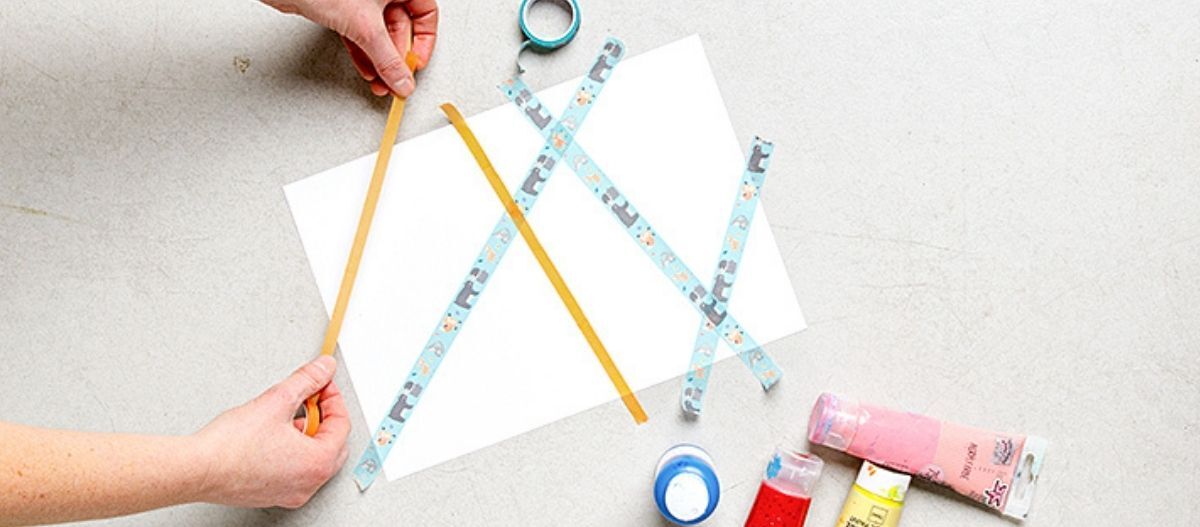 Ratgeber-Hund-DIY-Hunde-Kunst-schritt_1_1200x527 Zwei Hände befestigen Streifen aus Klebeband auf einem weißen Blatt Papier, das auf einer Arbeitsfläche liegt, um eine DIY-Hundekunst vorzubereiten; daneben liegen Farbtuben und eine Schere.