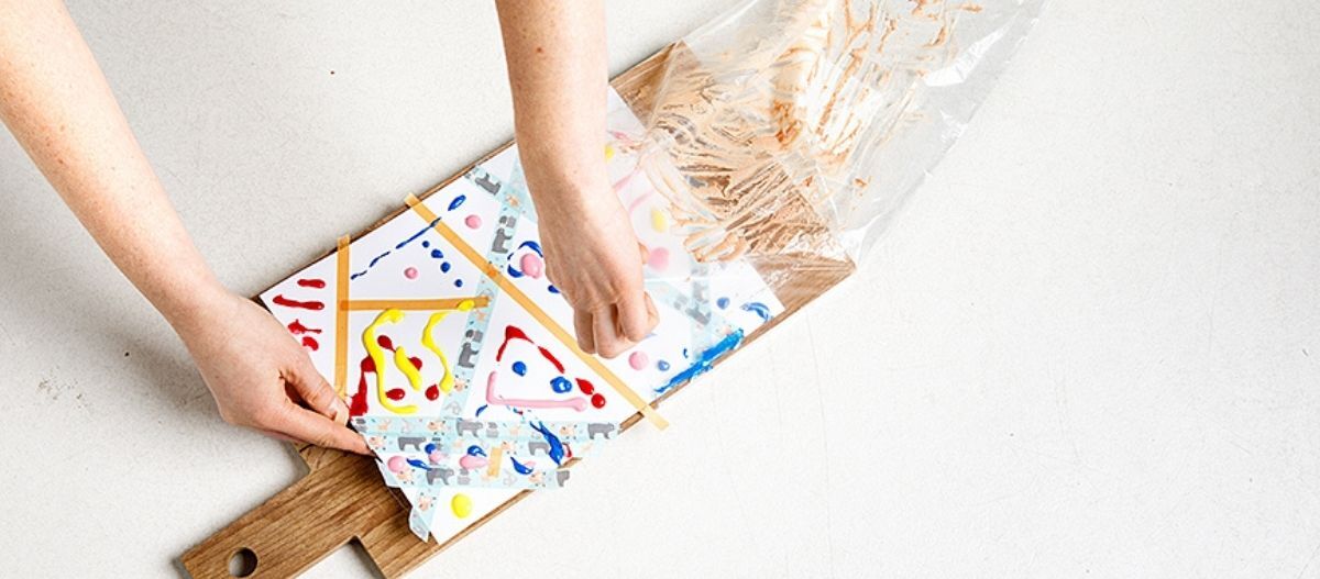 Ratgeber-Hund-DIY-Hunde-Kunst-schritt_4_1200x527 Zwei Hände legen ein Blatt Papier mit bunten Farbklecksen auf ein Holzbrett und bedecken es mit einer transparenten Plastikhülle für eine DIY-Hundekunst-Aktivität.