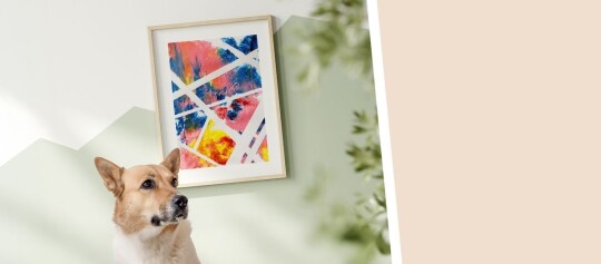 Ein Hund sitzt vor einer Wand, neben einem gerahmten, bunten abstrakten Kunstwerk mit geometrischen Linien in verschiedenen Farben.